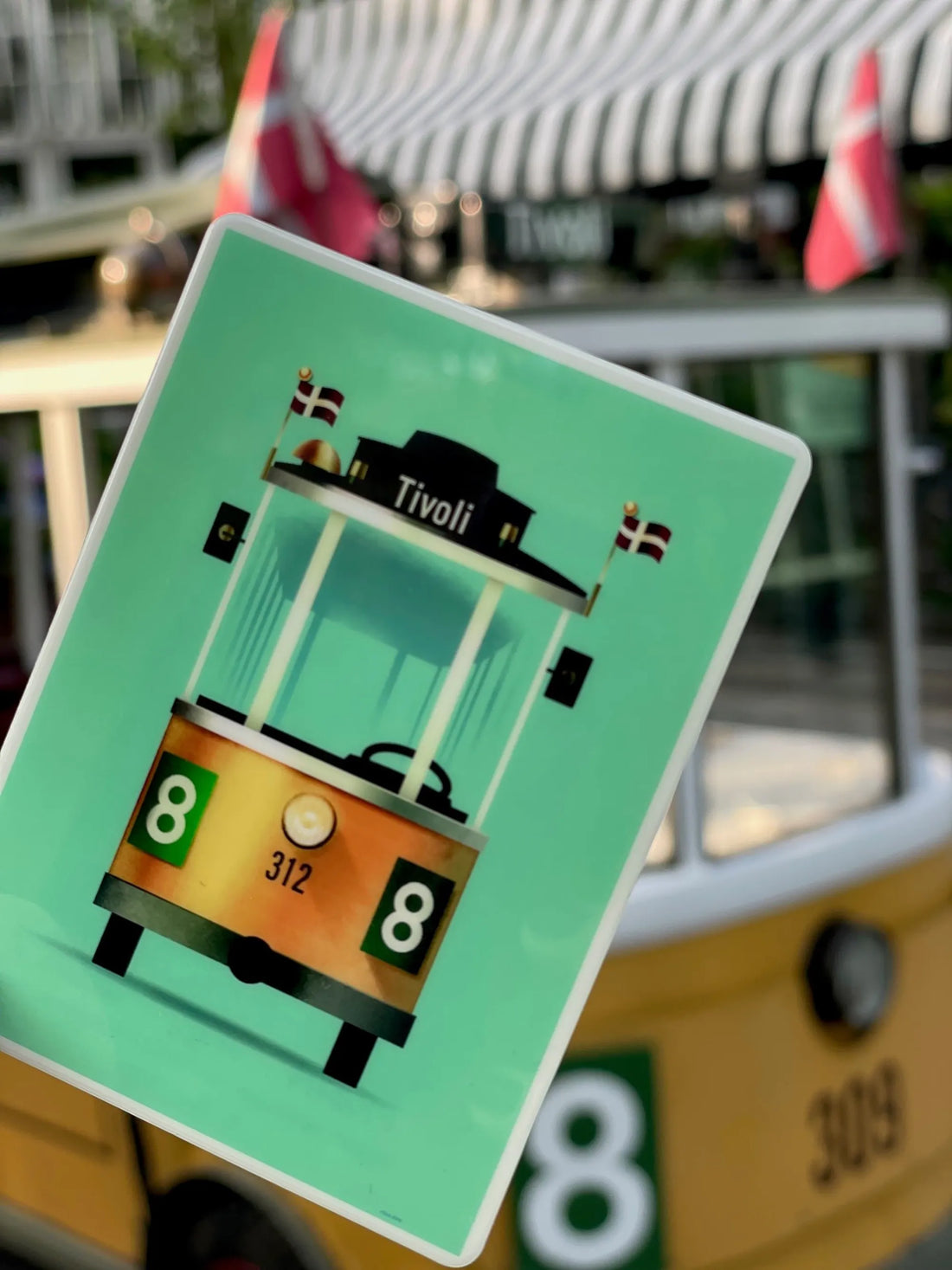 Linie 8 magnet med illustration af Tivolis ikoniske sporvogn designet af Mads Berg, fotograferet blandt grønne blade i Tivoli.