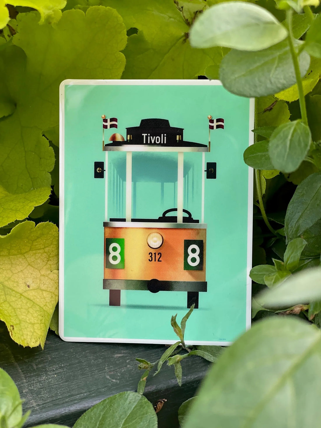 Linie 8 magnet med illustration af Tivolis ikoniske sporvogn designet af Mads Berg, fotograferet blandt grønne blade i Tivoli.