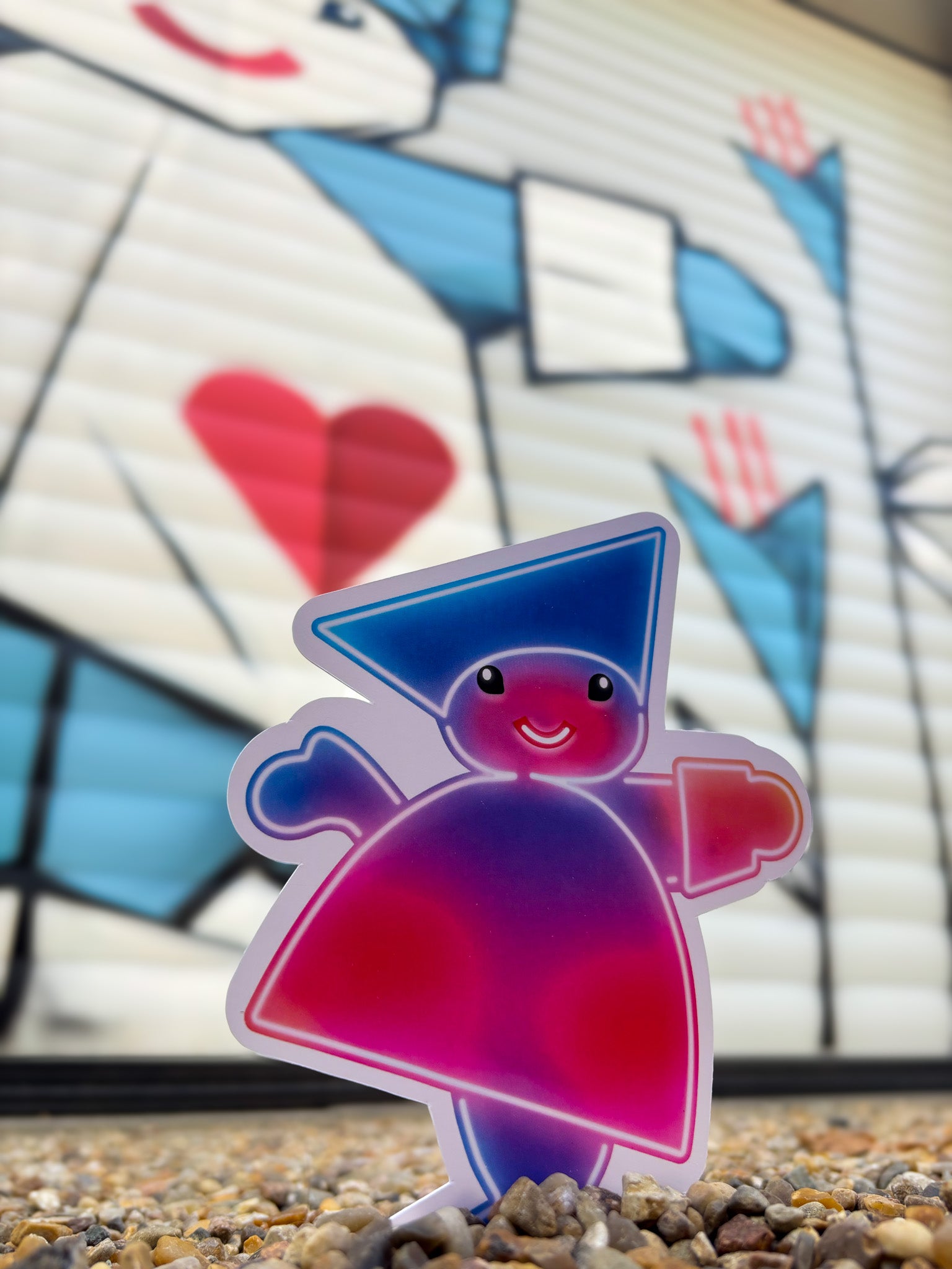 Little Tivoli Hikari Pjerrot-postkort med neoninspireret motiv placeret foran en stor illustration af Tivolis ikoniske Pjerrot. Postkortets blå og pink farver fremhæver forbindelsen til Hikaris magiske univers.