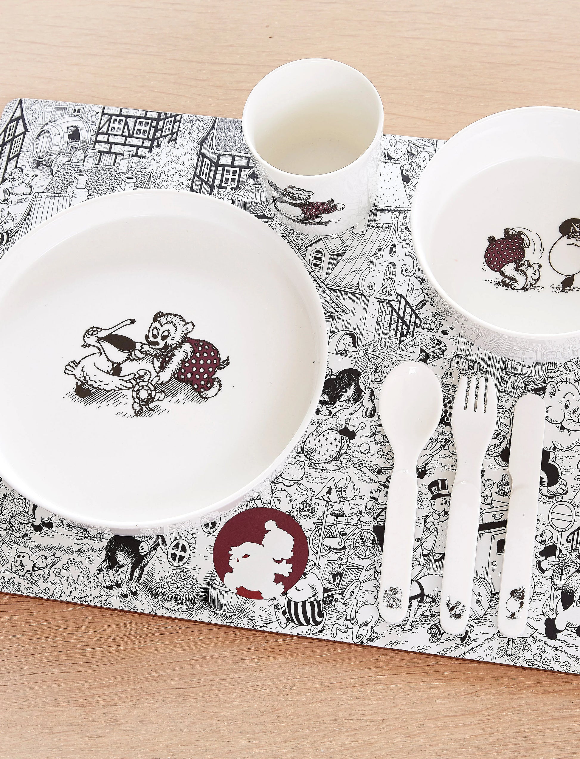 A children's tableware set arranged on a patterned placemat over a light wood surface, including a large white plate, a bowl, a cup, a spoon, a fork, and a knife, all printed with black-and-white cartoon illustrations and a small red accent. Rasmus Klump is the main character.