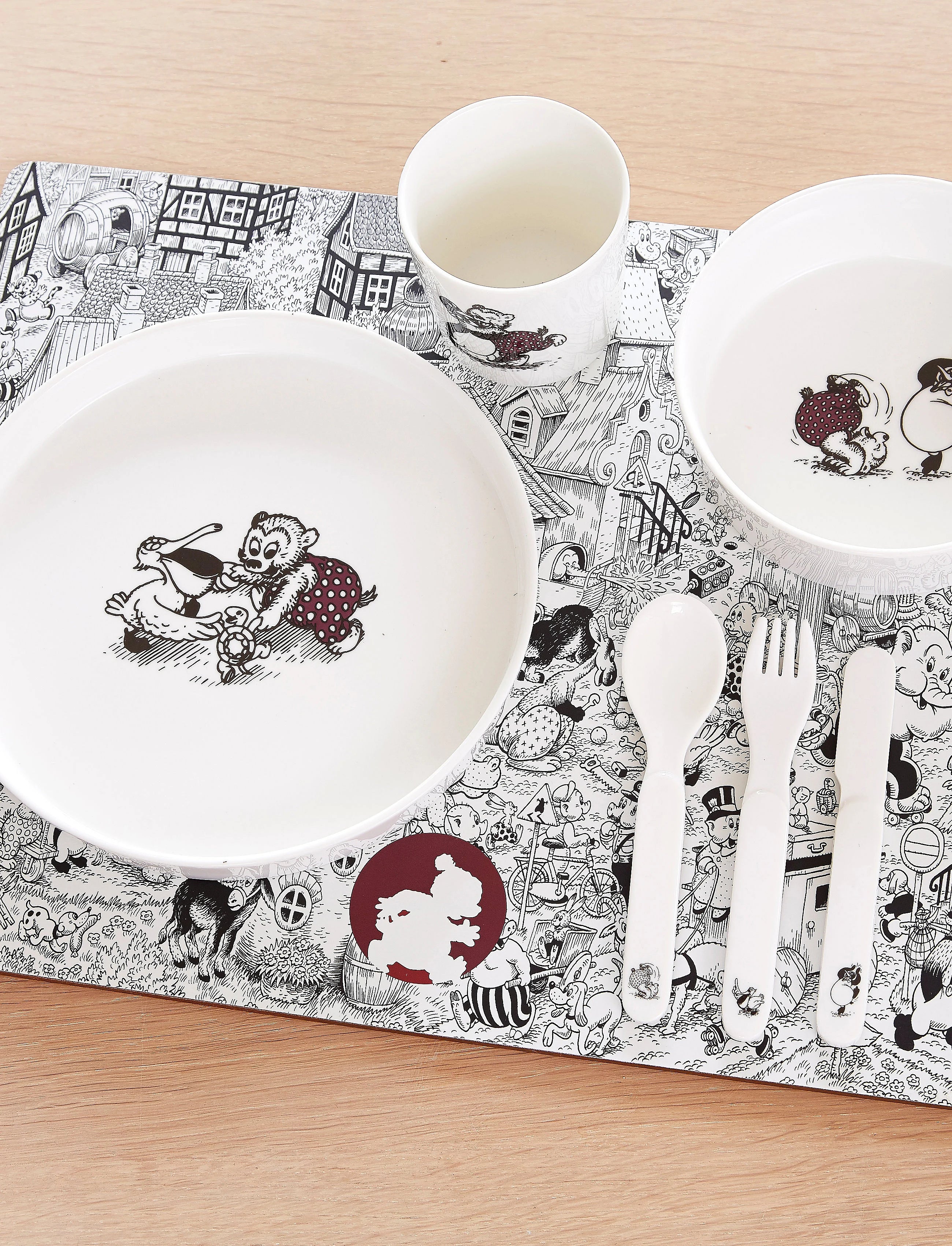 A children's tableware set arranged on a patterned placemat over a light wood surface, including a large white plate, a bowl, a cup, a spoon, a fork, and a knife, all printed with black-and-white cartoon illustrations and a small red accent. Rasmus Klump is the main character.