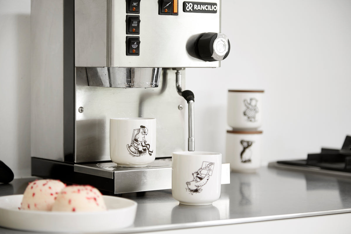 Rasmus Klump krus i porcelæn med klassiske illustrationer af Rasmus Klump-universets figurer ved siden af en espressomaskine