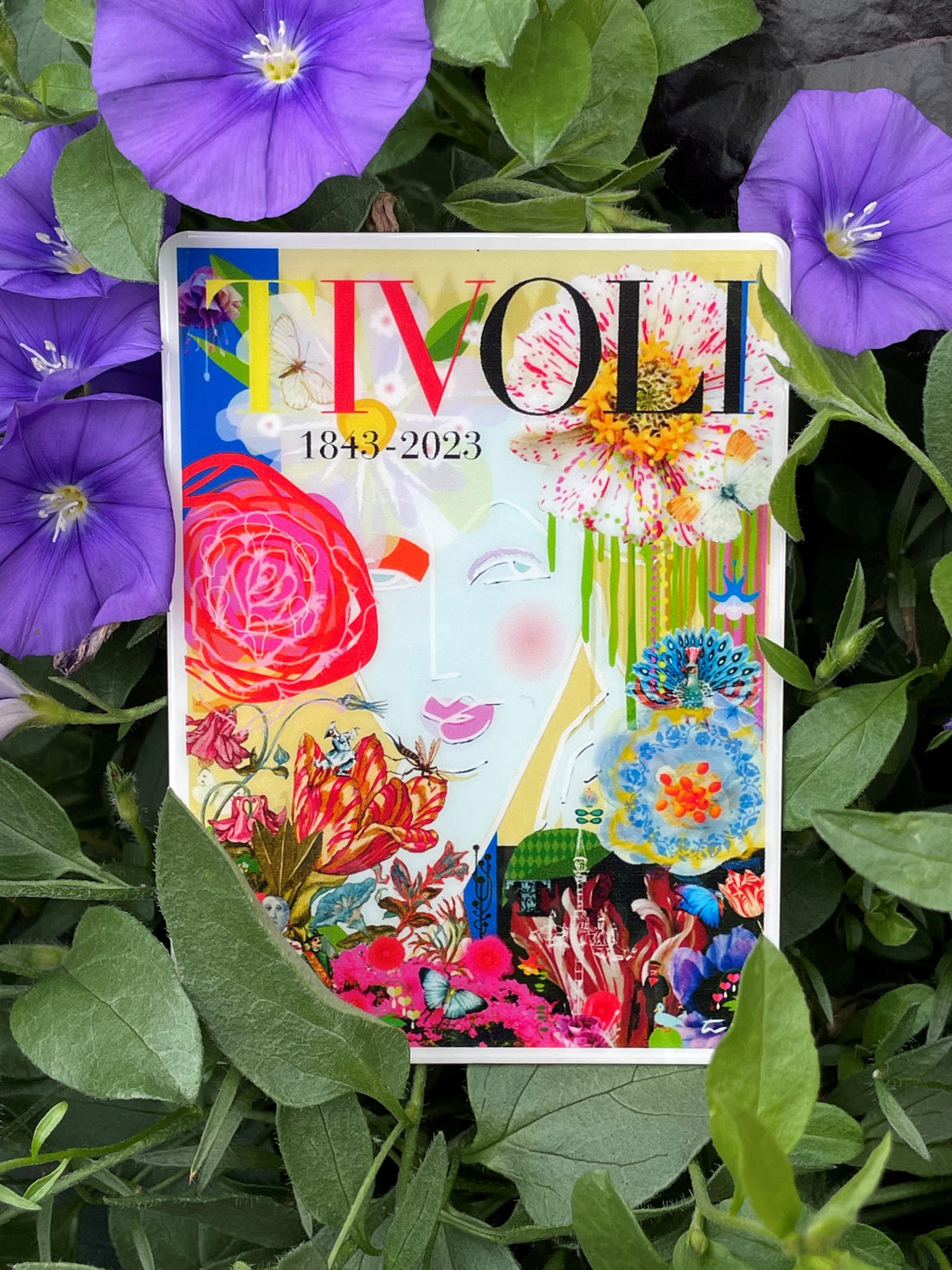 Tivoli magnet med farverigt jubilæumsmotiv skabt i anledning af Tivolis 180-års jubilæum 1843-2023. Designet viser blomster, sommerfugle, en påfugl, Tivoli-vartegn og en stiliseret ansigtssilhuet samlet i en kunstnerisk collage med teksten "TIVOLI 1843-2023" øverst.