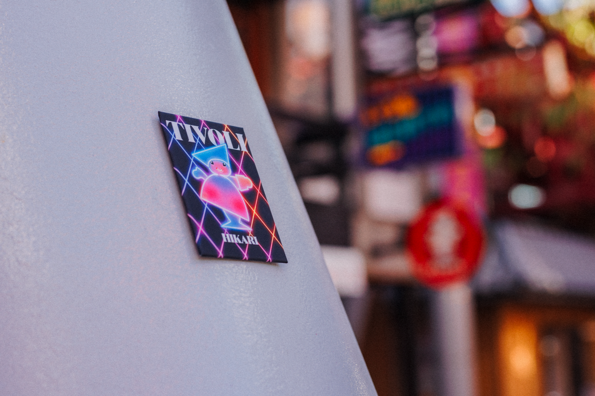 Tivoli HIKARI magnet badge med farverigt neoninspireret design monteret på en lys overflade. Motivet viser en stiliseret figur omgivet af lysende geometriske linjer og Tivoli-logo, mens baggrunden er sløret med farverige tivolilys.