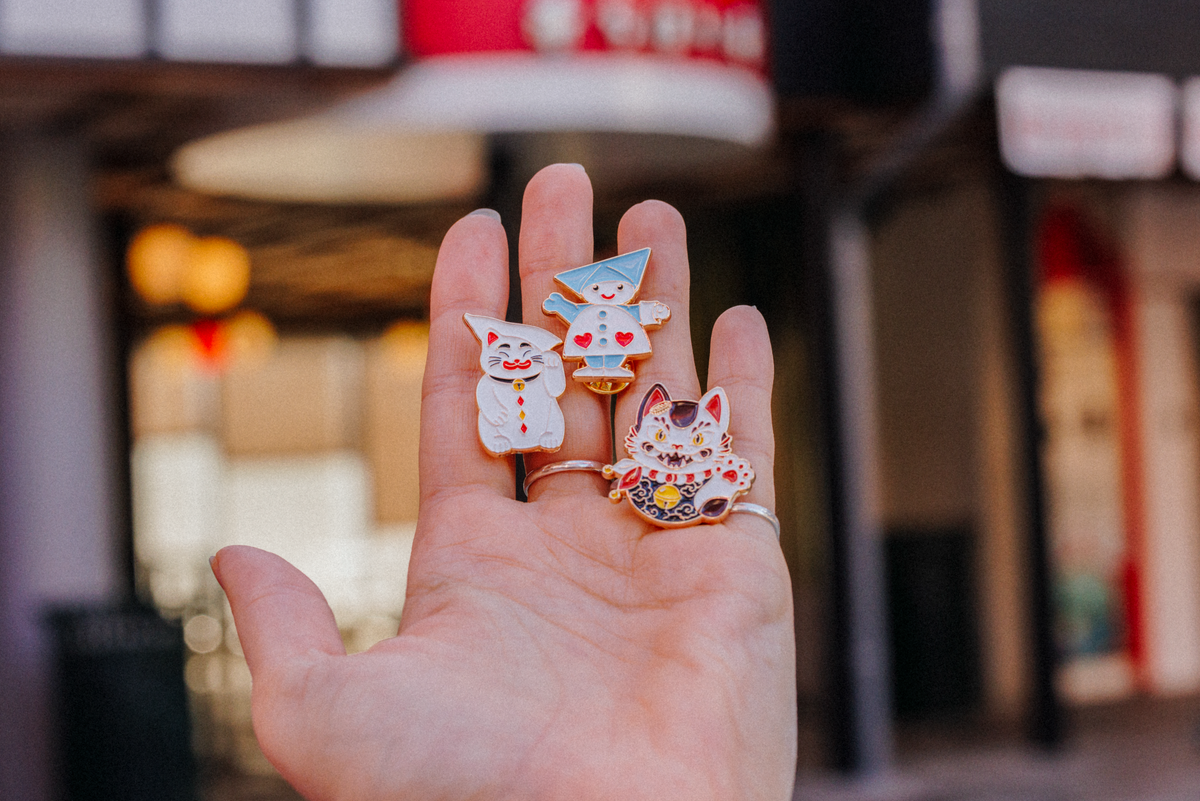 Tre Tivoli Hikari pins i emalje med farverige figurer inspireret af Hikari-universet. Motiverne viser en kat, en geometrisk figur og en lykkekat i neoninspirerede farver og detaljer.