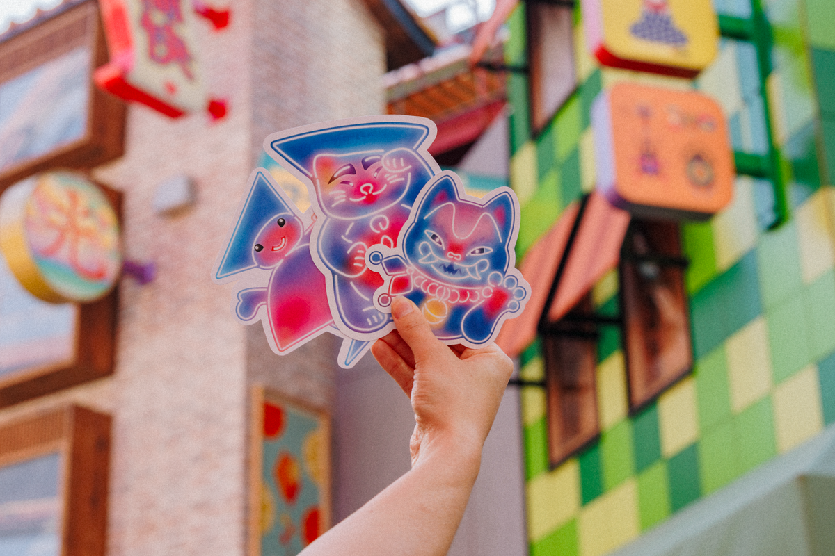 Tivoli Hikari post kort med tre farverige neonfigurer i lilla, blå og pink nuancer. Klistermærket holdes op foran den farverige Hikari-attraktion i Tivoli og viser det karakteristiske lysende design fra attraktionen. 