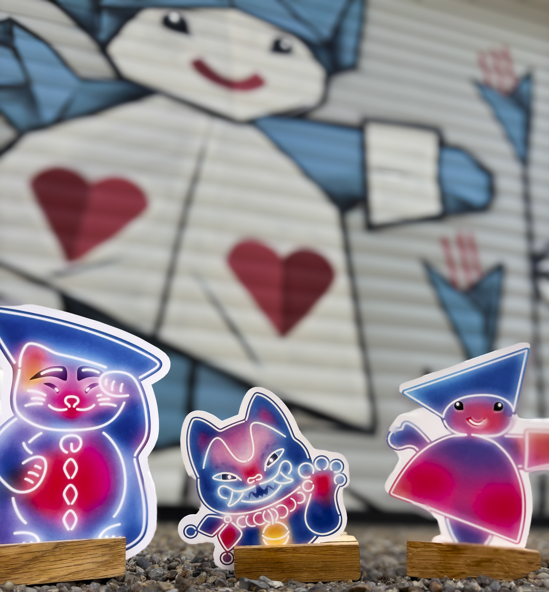 Farverige neoninspirerede figurer i blå, pink og lilla står i forgrunden, herunder en kattefigur og geometriske karakterer. I baggrunden ses et stort gadekunstmaleri på en rulleskodde med en stiliseret figur dekoreret med røde hjerter, blå detaljer og pilemotiver. Motivet skaber en kreativ og kunstnerisk stemning i et urbant miljø.