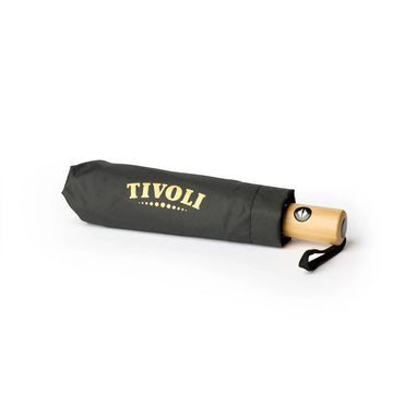Sammenfoldet Tivoli paraply med sort betræk, guldfarvet TIVOLI-logo og håndtag i lyst træ. Den kompakte paraply har håndledsrem og er designet til nem opbevaring i taske eller rygsæk.