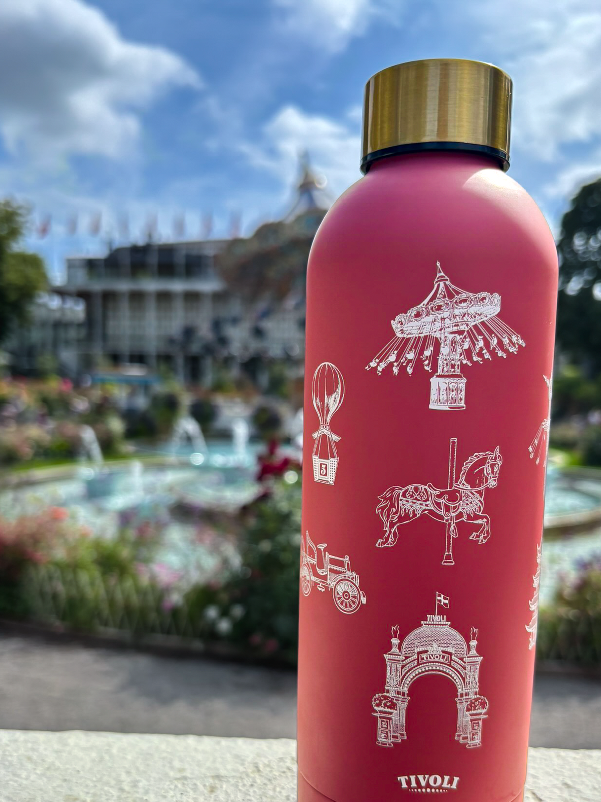 Rosa Tivoli termoflaske med guldfarvet skruelåg og hvide illustrationer af ikoniske Tivoli-motiver, herunder Hovedindgangen, karusselhest, Ballongyngen og hestevogn. Termoflasken står foran Tivolis springvand og Koncertsalen i baggrunden.