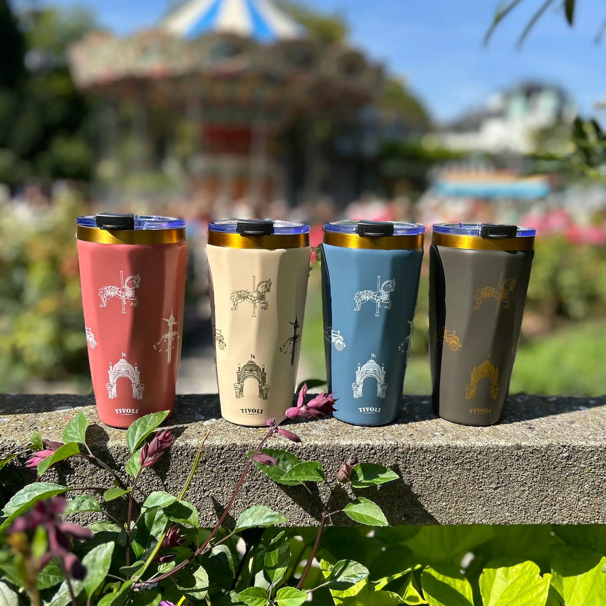 Tivoli To-Go termokrus i rosa, beige, blå og grå med guldfarvede detaljer og ikoniske Tivoli-motiver. Krusene står udendørs i Tivoli med Karussellen og blomster sløret i baggrunden.